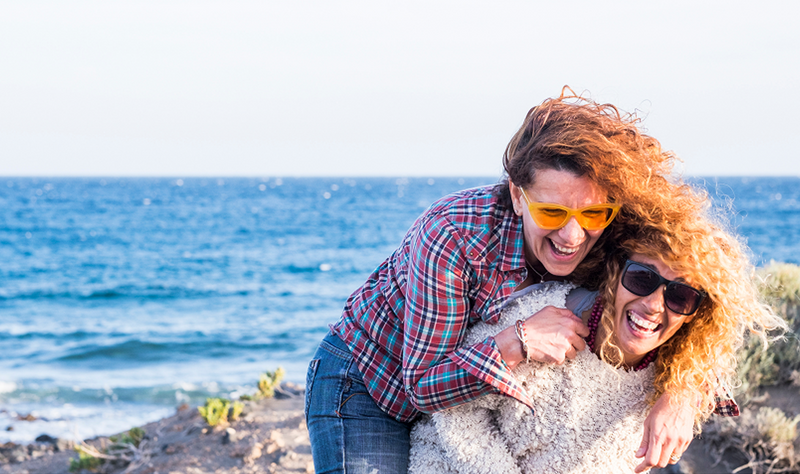 Two women with curly hair and sunglasses laugh and embrace on a sandy coastal cliff overlooking the blue ocean, representing the vitality and joy of healthy aging.